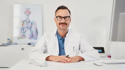 Zuversichtlich-aussehender Arzt an Schreibtisch sitzend in Arztzimmerumgebung mit Vivatmo pro im Hintergrund