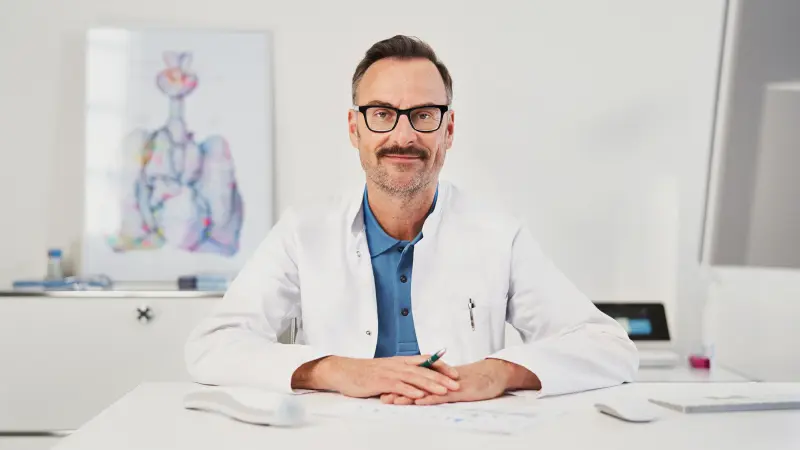 Zuversichtlich-aussehender Arzt an Schreibtisch sitzend in Arztzimmerumgebung mit Vivatmo pro im Hintergrund
